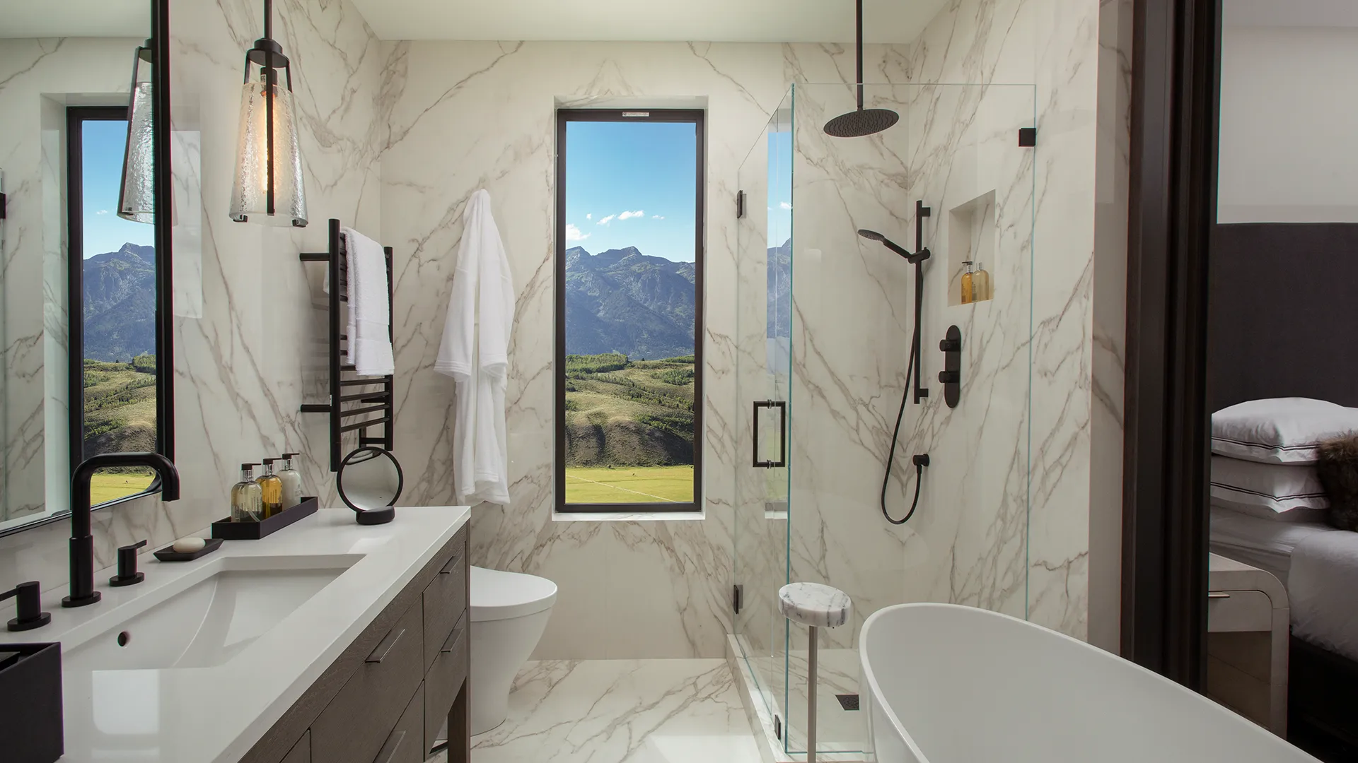 Luxury bathroom with marble finishes, modern fixtures, and a view of mountains through a large window, featuring a freestanding tub and glass shower.