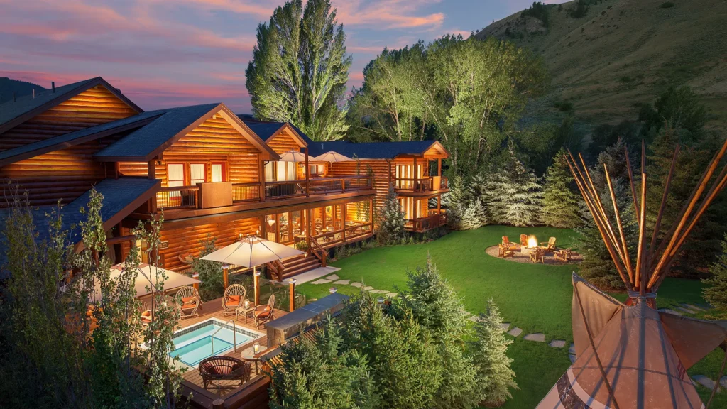 Rustic luxury resort with wooden architecture, outdoor seating, pool, and fire pit, surrounded by trees and mountains, highlighting serene and inviting atmosphere for guests.